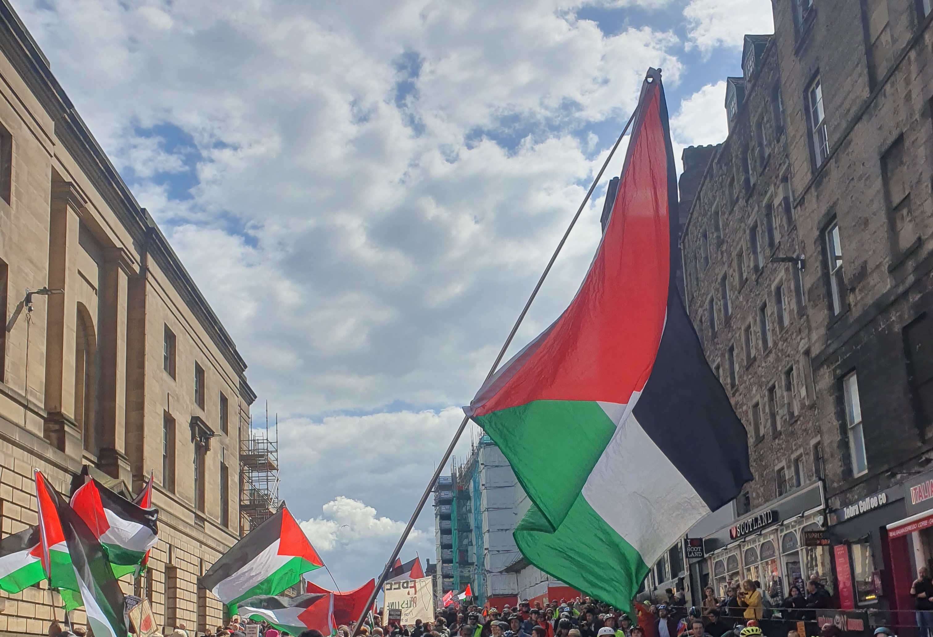 Palestine flags are held aloft during a protest. Buildings and a blue cloudy sky are in the background