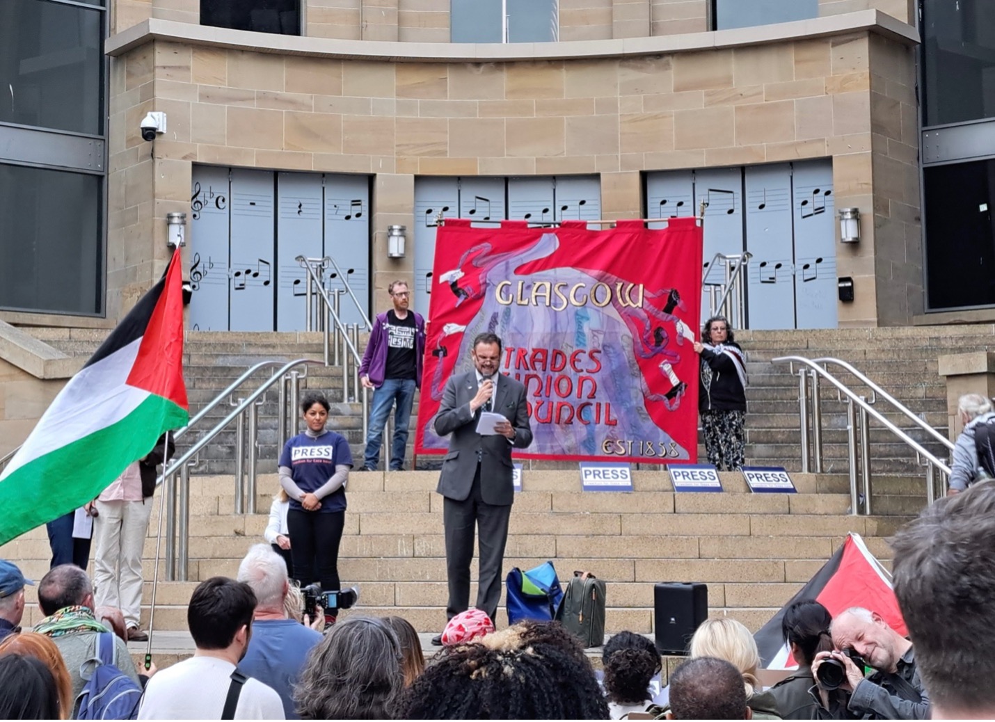 Speaker standing on steps in front of crowd. Building behind speaker with red banner in background. Crowd in foreground waving Palestine flags 